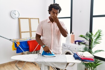 African man with curly hair ironing clothes at home smelling something stinky and disgusting,...