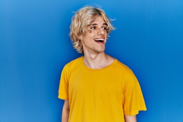 Young modern man standing over blue background looking away to side with smile on face, natural expression. laughing confident.