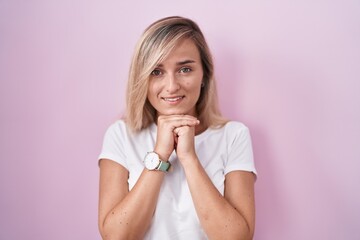Young blonde woman standing over pink background laughing nervous and excited with hands on chin...
