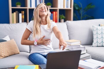 Young blonde woman studying using computer laptop at home laughing at you, pointing finger to the...