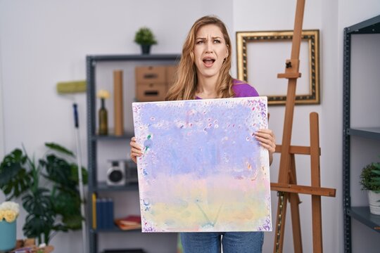 Beautiful blonde woman holding painting canvas angry and mad screaming frustrated and furious, shouting with anger. rage and aggressive concept.