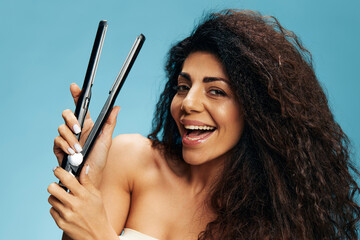 Beautiful enjoyed Latin curly woman using hair straightener, looking at camera, posing isolated on...