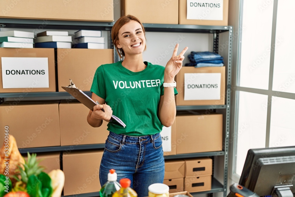 Canvas Prints Young brunette woman wearing volunteer t shirt at donations stand smiling with happy face winking at the camera doing victory sign with fingers. number two.