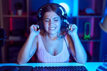 Young blonde woman playing video games wearing headphones covering ears with fingers with annoyed expression for the noise of loud music. deaf concept.