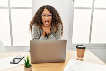 Beautiful hispanic business woman sitting on desk at office working with laptop shouting and suffocate because painful strangle. health problem. asphyxiate and suicide concept.