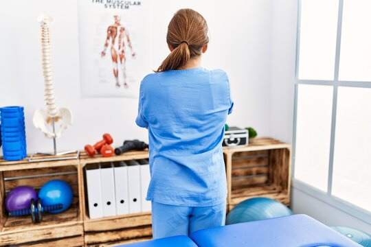Middle age hispanic physiotherapist woman working at pain recovery clinic standing backwards looking away with crossed arms