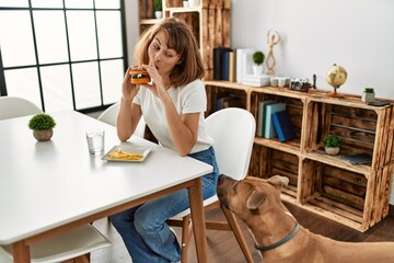 Young caucasian woman eating classical hamburger sitting on table at home