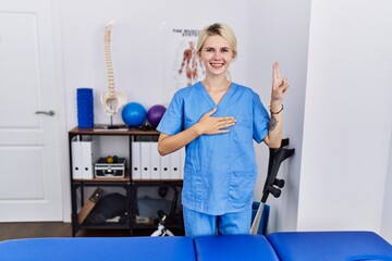 Young physiotherapist woman working at pain recovery clinic smiling swearing with hand on chest and fingers up, making a loyalty promise oath