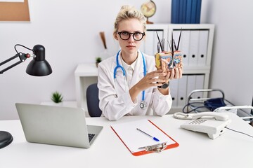 Young doctor woman holding model of human anatomical skin and hair at the clinic thinking attitude and sober expression looking self confident