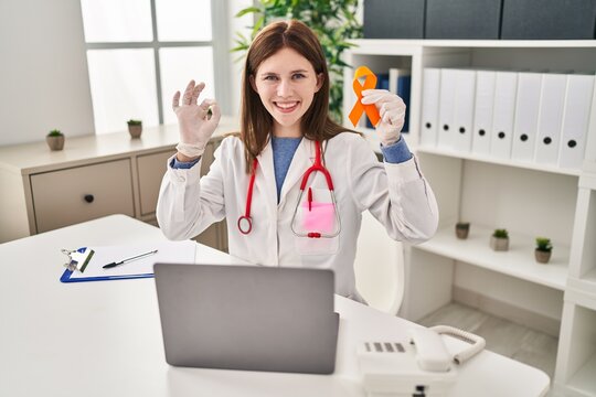 Young doctor woman holding awareness orange ribbon doing ok sign with fingers, smiling friendly gesturing excellent symbol