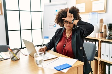 African american woman with afro hair working at the office wearing operator headset making fun of people with fingers on forehead doing loser gesture mocking and insulting. - obrazy, fototapety, plakaty