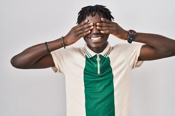 African man with dreadlocks standing over isolated background covering eyes with hands smiling...