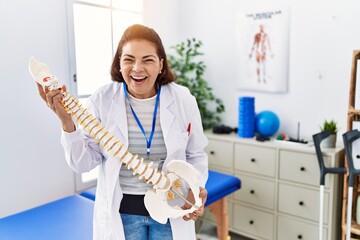 Middle age hispanic physiotherapy woman holding anatomical model of spinal column smiling and...