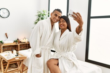 Young latin couple wearing bathrobe making selfie by the smartphone at beauty center.