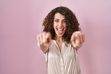 Hispanic woman with curly hair standing over pink background pointing to you and the camera with fingers, smiling positive and cheerful