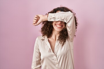 Naklejka premium Hispanic woman with curly hair standing over pink background covering eyes with arm smiling cheerful and funny. blind concept.