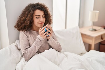 Young hispanic woman drinking cup of coffee sitting on bed at bedroom