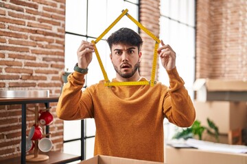 Hispanic man with beard moving to a new home holding ruler in shock face, looking skeptical and...