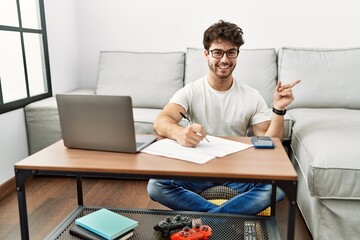 Hispanic man doing papers at home with a big smile on face, pointing with hand finger to the side looking at the camera.