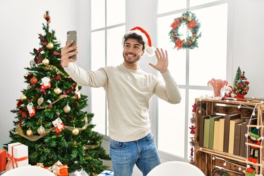 Young hispanic man wearing christmas hat making selfie by the smartphone at home.