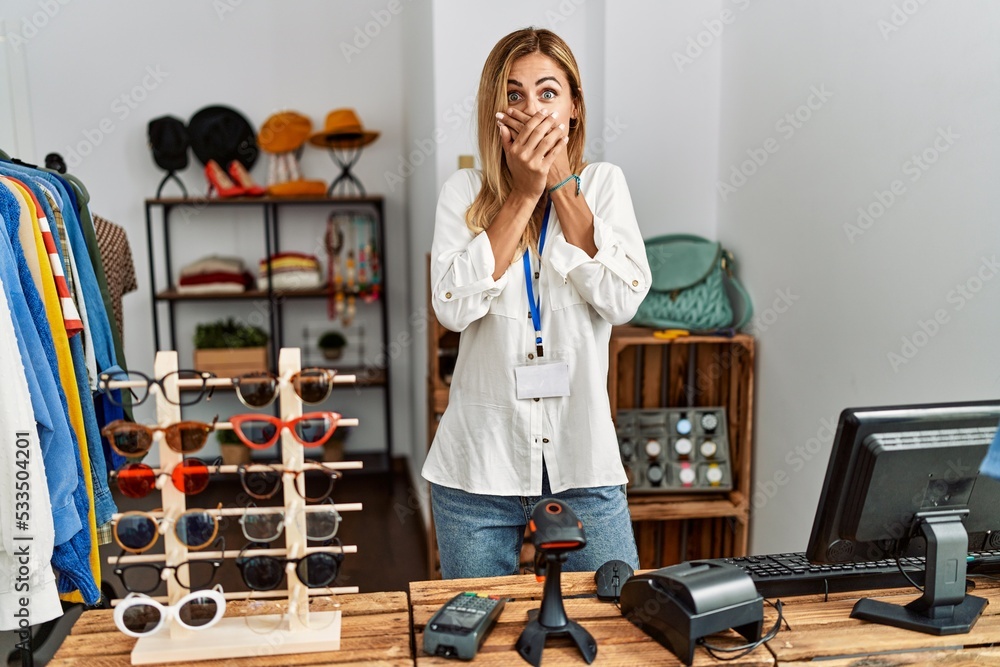 Poster blonde beautiful young woman working as manager at retail boutique shocked covering mouth with hands