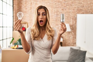 Young blonde woman holding led lightbulb and incandescent bulb in shock face, looking skeptical and...