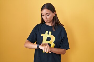 Young hispanic woman wearing bitcoin t shirt checking the time on wrist watch, relaxed and confident