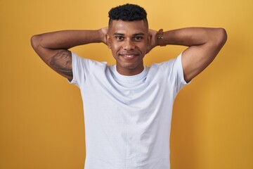 Fototapeta na wymiar Young hispanic man standing over yellow background relaxing and stretching, arms and hands behind head and neck smiling happy