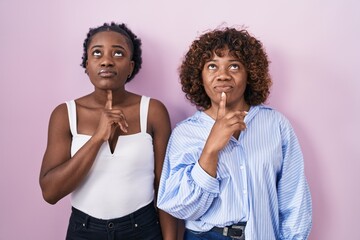 Two african women standing over pink background thinking concentrated about doubt with finger on chin and looking up wondering