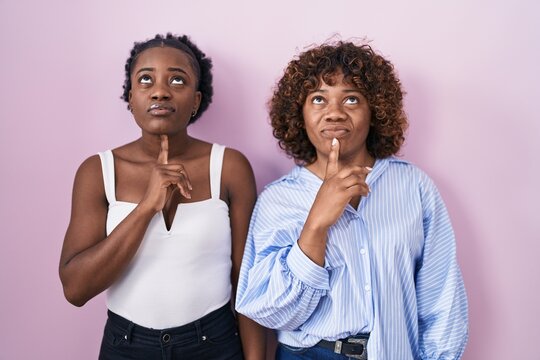 Two African Women Standing Over Pink Background Thinking Concentrated About Doubt With Finger On Chin And Looking Up Wondering