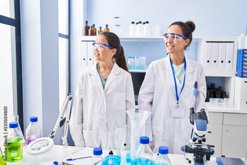 Sticker young mother and daughter at scientist laboratory looking away to side with smile on face, natural e