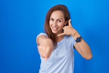 Brunette woman standing over blue background smiling doing talking on the telephone gesture and...