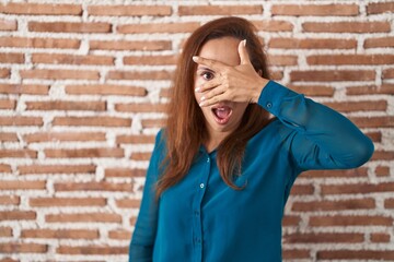 Brunette woman standing over bricks wall peeking in shock covering face and eyes with hand, looking through fingers with embarrassed expression.