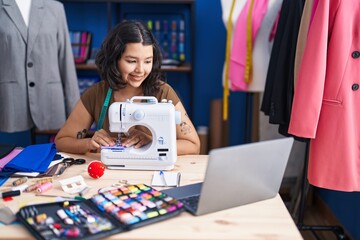 Fototapeta premium Young woman tailor using sewing machine and laptop at sewing studio