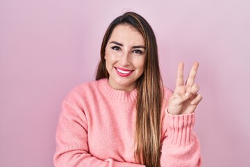 Young hispanic woman standing over pink background smiling with happy face winking at the camera doing victory sign with fingers. number two.