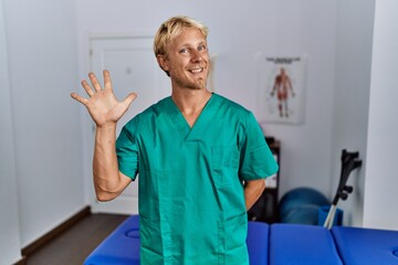 Young blond man wearing physiotherapist uniform standing at clinic showing and pointing up with fingers number five while smiling confident and happy.