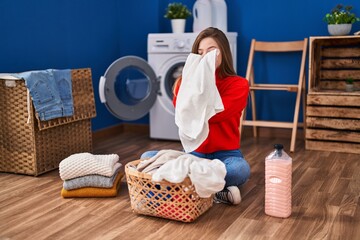 Young blonde woman smelling clothes sitting on floor at laundry room