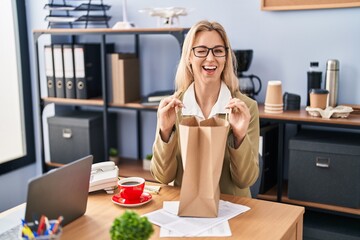 Young caucasian woman working at the office holding paper bag winking looking at the camera with...