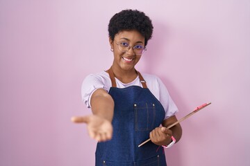 Young african american woman wearing professional waitress apron holding clipboard smiling cheerful offering palm hand giving assistance and acceptance.