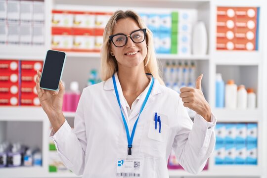 Young blonde woman working at pharmacy drugstore showing smartphone screen smiling happy and positive, thumb up doing excellent and approval sign