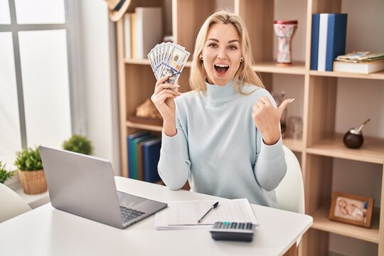 Young caucasian woman using laptop holding dollars banknotes pointing thumb up to the side smiling happy with open mouth