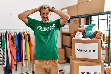 Middle age hispanic man wearing volunteer t shirt at donations stand crazy and scared with hands on...