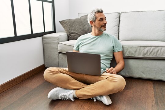 Middle Age Hispanic Man Using Laptop Sitting On The Floor At The Living Room Looking To Side, Relax Profile Pose With Natural Face And Confident Smile.