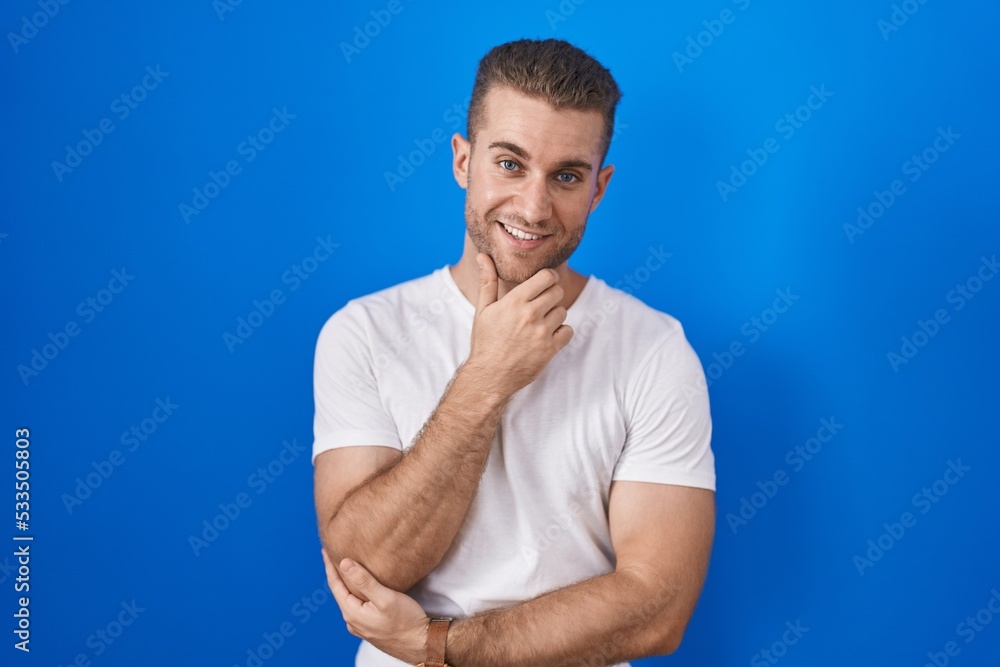 Sticker young caucasian man standing over blue background looking confident at the camera smiling with cross