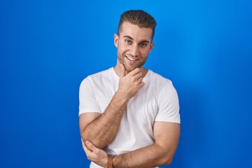 Young caucasian man standing over blue background looking confident at the camera smiling with crossed arms and hand raised on chin. thinking positive.