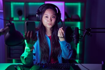 Young asian woman playing video games with smartphone doing money gesture with hands, asking for salary payment, millionaire business