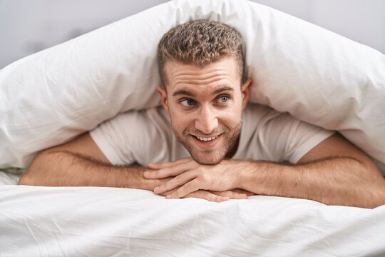 Young caucasian man lying on bed covering with bedsheet at bedroom