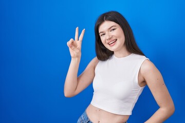 Fototapeta premium Young caucasian woman standing over blue background smiling looking to the camera showing fingers doing victory sign. number two.