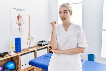 Obraz premium Young caucasian woman working at pain recovery clinic smiling with happy face looking and pointing to the side with thumb up.