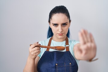 Young modern girl with blue hair wearing cook apron holding spoon with open hand doing stop sign with serious and confident expression, defense gesture